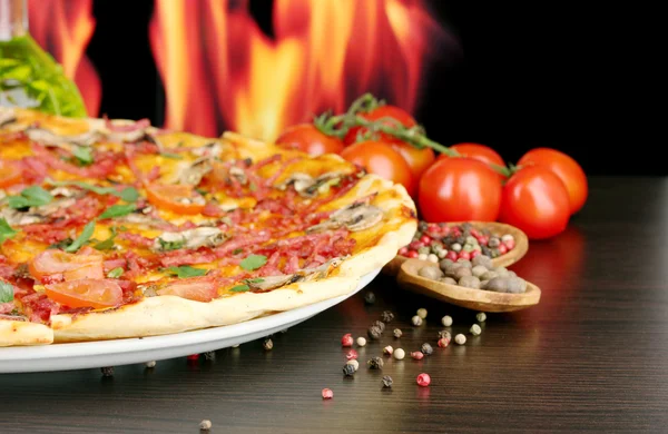 Delicious pizza and tomatoes on wooden table on flame background — Stock Photo, Image