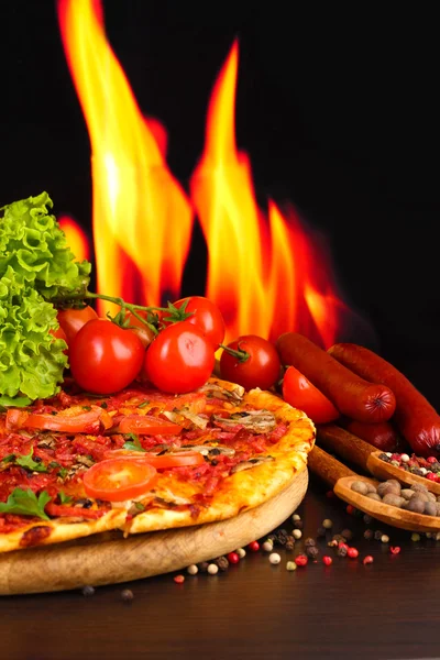 Deliciosa pizza, salami, tomates y especias en mesa de madera sobre fondo de llama —  Fotos de Stock