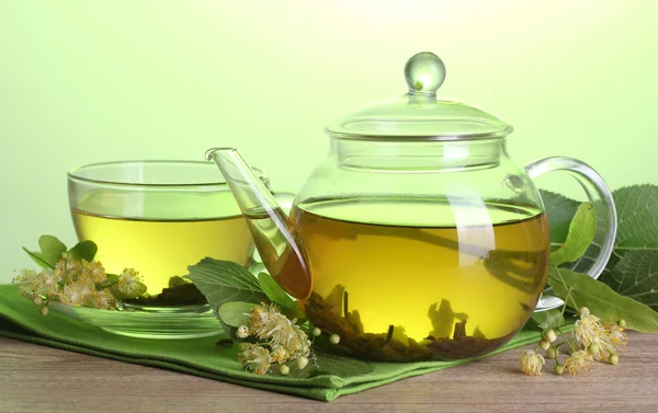 Tekanna och kopp med linden te och blommor på träbord på grön bakgrund — Stockfoto