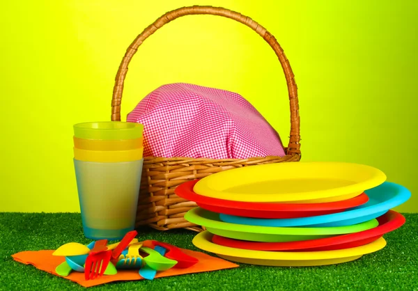 Bright plastic disposable tableware and picnic basket on the lawn on colorful background — Stock Photo, Image