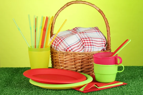 Plástico brilhante utensílios de mesa descartáveis e cesta de piquenique no gramado em fundo colorido — Fotografia de Stock