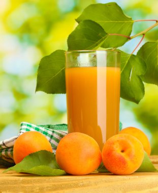 Glass of apricot juice and fresh apricots on wooden table on green background clipart