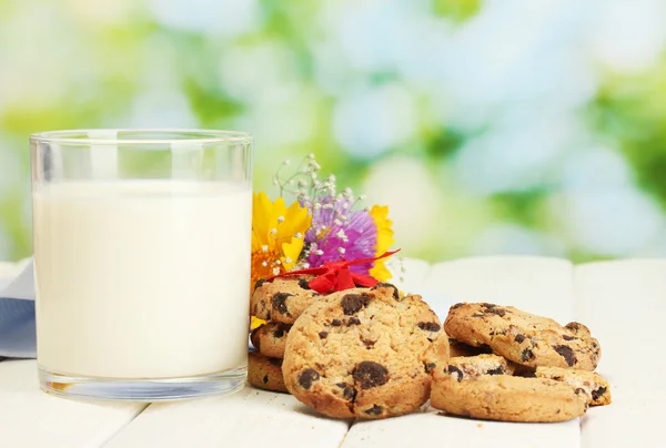 Склянка молока, шоколадне печиво з червоною стрічкою та польовими квітами на дерев'яному столі на зеленому фоні — стокове фото