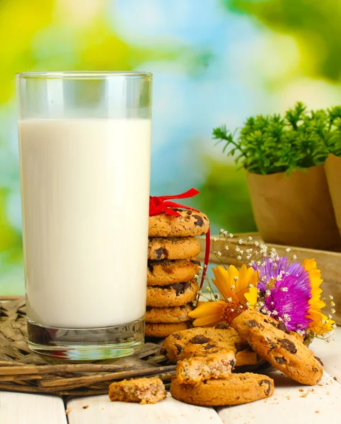 Склянка молока, шоколадне печиво з червоною стрічкою та польовими квітами на дерев'яному столі на зеленому фоні — стокове фото
