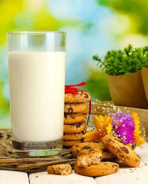 Glass of milk, chocolate chips cookies with red ribbon and wildflowers on wooden table on green background — Stock Photo, Image