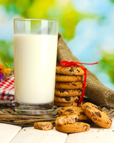 Glass of milk, chocolate chips cookies with red ribbon and wildflowers on wooden table on green background — Stock Photo, Image