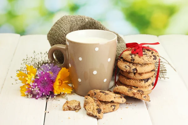 Tasse de lait, biscuits aux pépites de chocolat avec ruban rouge et fleurs sauvages sur table en bois sur fond vert — Photo