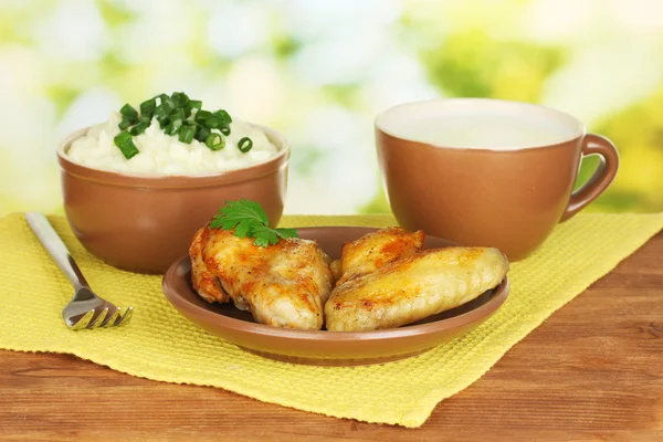 Kartoffelpüree in der Schüssel und gebratene Hühnerflügel im Teller und Tasse mit Milch auf bunter Serviette auf Holztisch in Großaufnahme — Stockfoto