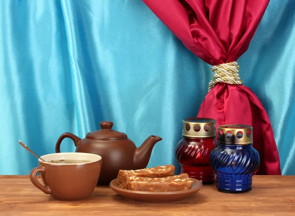 Чайник с чашкой и блюдцем со сладким шербетом на деревянном столе на фоне занавеса крупным планом — стоковое фото