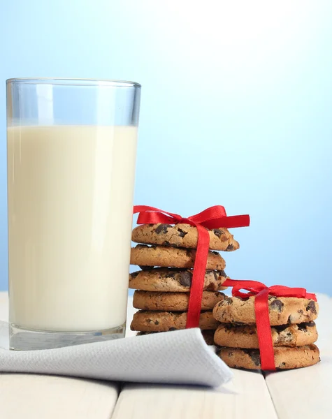 Glass of milk and chocolate chips cookies with red ribbon on wooden table on blue background — Stock Photo, Image