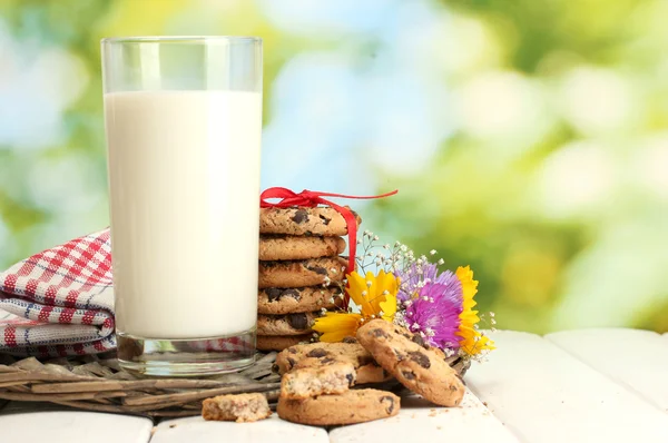 Szklanki mleka, czekoladowe żetony ciasteczka z czerwoną wstążką i kwiaty na drewnianym stole na zielonym tle — Zdjęcie stockowe
