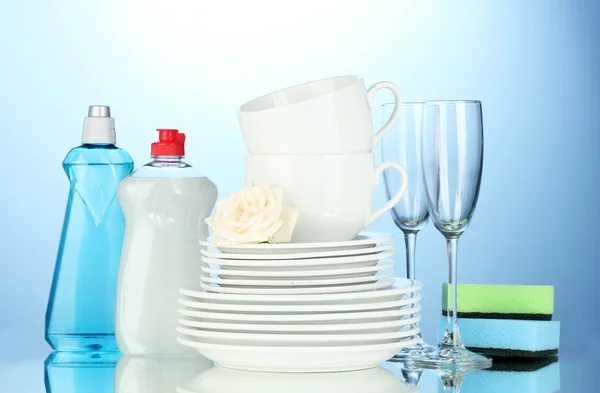 Порожні чисті тарілки, окуляри та чашки з посудомийною рідиною та губками на синьому фоні — стокове фото