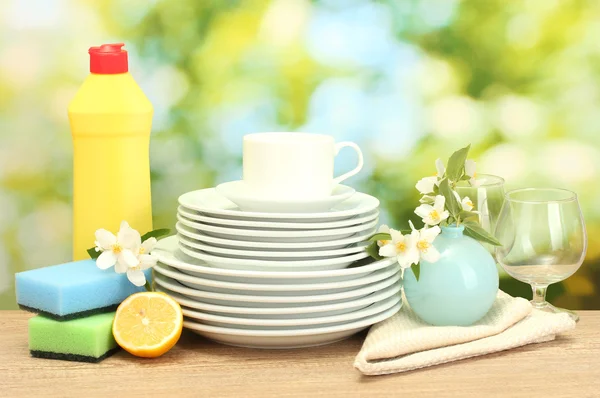 Platos limpios vacíos, vasos y tazas con líquido lavavajillas, esponjas y limón sobre mesa de madera sobre fondo verde — Foto de Stock