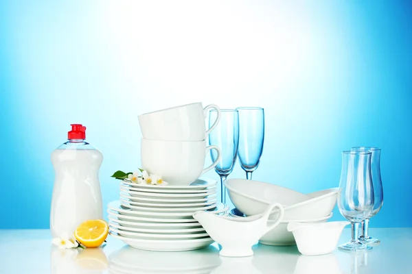 Platos limpios vacíos, vasos y tazas con líquido lavavajillas y limón sobre fondo azul — Foto de Stock