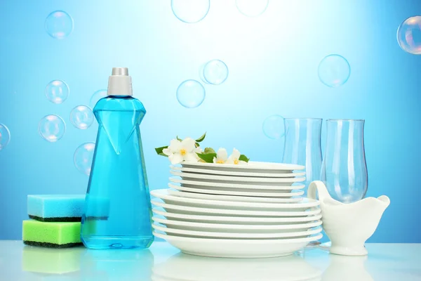 Farfurii curate goale și ochelari cu lichid de spălat vase, bureți și flori pe fundal albastru — Fotografie, imagine de stoc