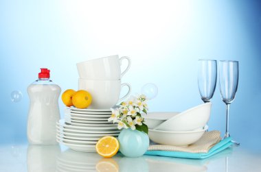 Empty clean plates, glasses and cups with dishwashing liquid and lemon on blue background clipart