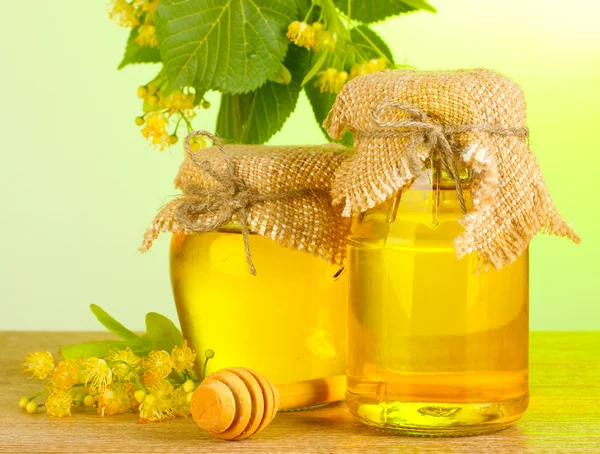 Jars with linden honey and flowers on wooden table on green background