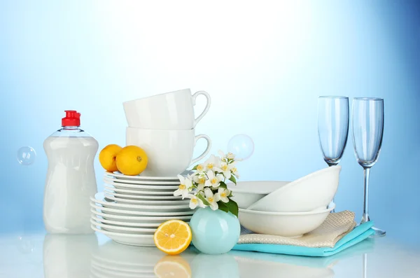 Platos limpios vacíos, vasos y tazas con líquido lavavajillas y limón sobre fondo azul — Foto de Stock