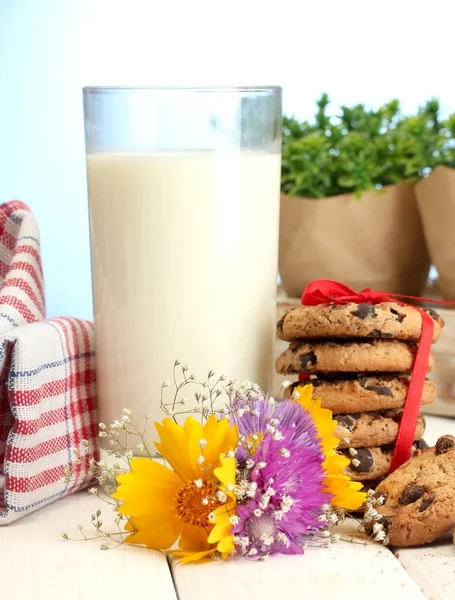 Glas Milch, Schokoladenkekse mit roter Schleife und Wildblumen auf Holztisch auf blauem Hintergrund — Stockfoto