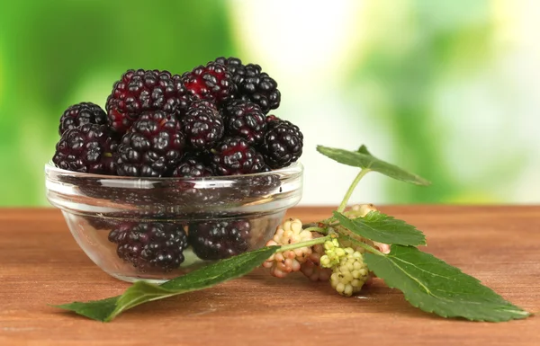 Transparent bowl with ripe mulberries on wooden table on bright green background close-up — Stock Photo, Image