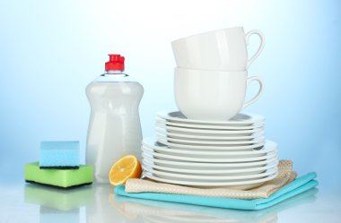 Empty clean plates and cups with dishwashing liquid, sponges and lemon on blue background clipart