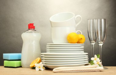 Empty clean plates, glasses and cups with dishwashing liquid, sponges and lemon on wooden table on grey background clipart