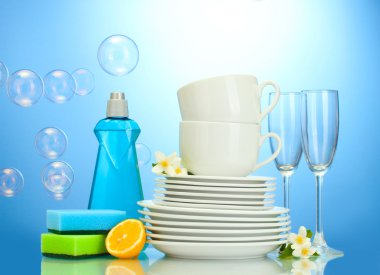 Empty clean plates, glasses and cups with dishwashing liquid, sponges and lemon on blue background clipart