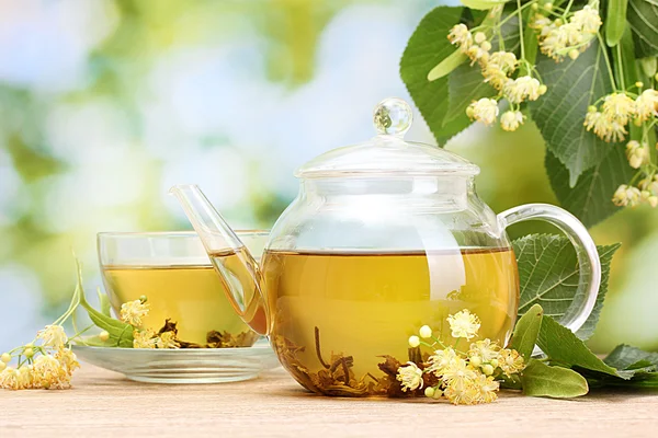 Tetera y taza con té de tilo y flores en la mesa de madera en el jardín — Foto de Stock
