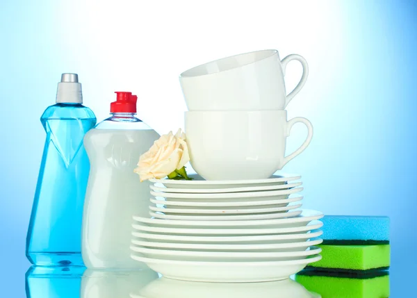 Platos y tazas limpias vacías con líquido lavavajillas y esponjas sobre fondo azul —  Fotos de Stock