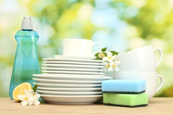 Platos y tazas limpias vacías con líquido lavavajillas, esponjas y limón sobre una mesa de madera sobre fondo verde — Foto de Stock