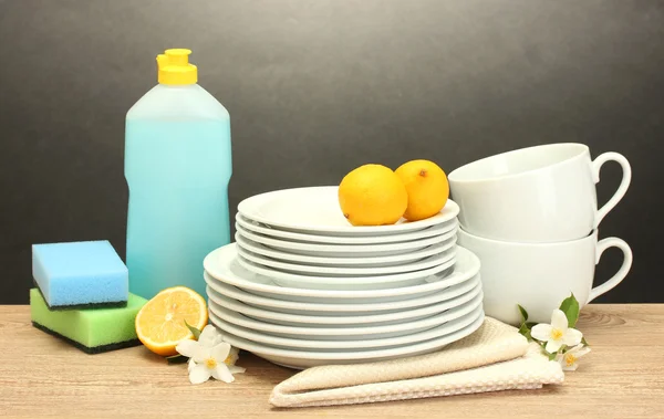 Piatti puliti vuoti, bicchieri e tazze con detersivo per piatti, spugne e limone su tavolo in legno su fondo grigio — Foto Stock