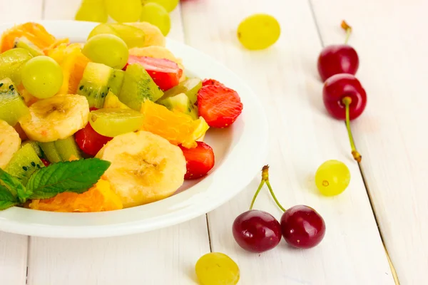 Fresh fruits salad on plate and berries on white wooden table — Stock Photo, Image