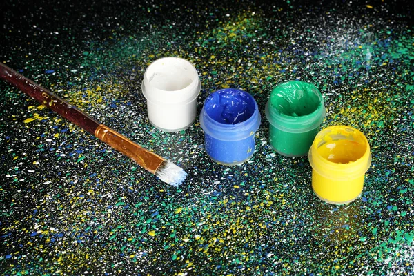 stock image Jars with colorful gouache and brush on black background, spattered with colorful paint close-up