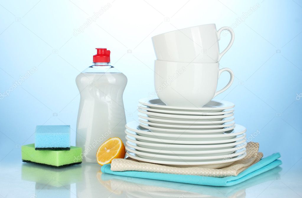 Empty clean plates and cups with dishwashing liquid, sponges and lemon on blue background