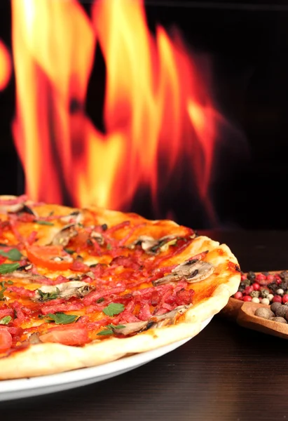 stock image Delicious pizza with vegetables and salami on wooden table on flame background