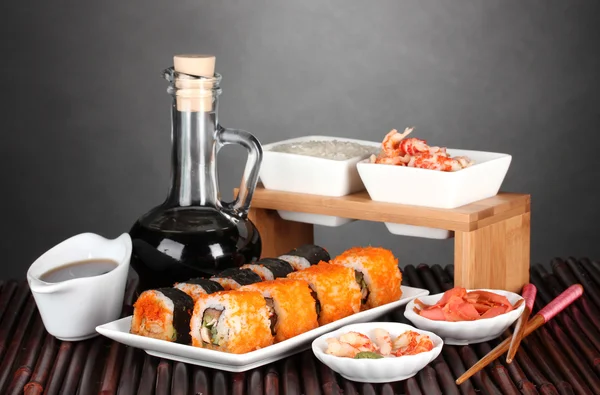 Delicious sushi on plate, chopsticks, soy sauce, fish and shrimps on bamboo mat on gray background — Stock Photo, Image