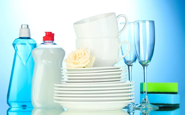 Empty clean plates, glasses and cups with dishwashing liquid and sponges on blue background