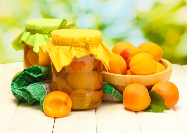 stock image Canned apricots in a jars and ripe apricots in bowl on wooden table on green background