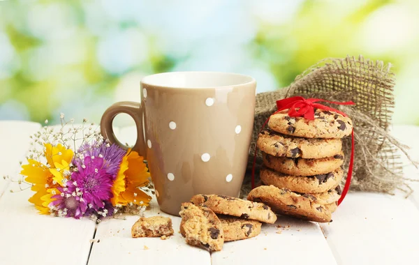 Tazza di latte, gocce di cioccolato biscotti con nastro rosso e fiori di campo su tavolo di legno su sfondo verde — Foto Stock