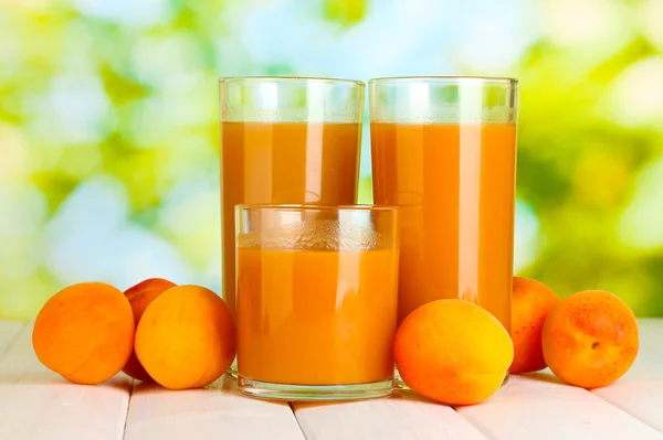 Glasses of apricot juice and fresh apricots on white wooden table on green background — Stock Photo, Image