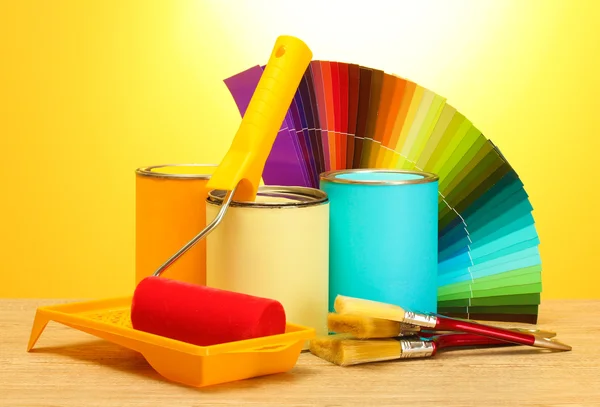 Blechdosen mit Farbe, Rolle, Pinsel und heller Farbpalette auf Holztisch auf gelbem Hintergrund — Stockfoto