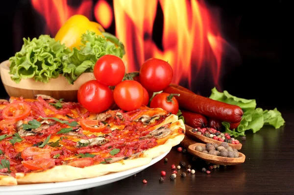Heerlijke pizza, salami, groenten en kruiden op houten tafel op vlam achtergrond — Stockfoto