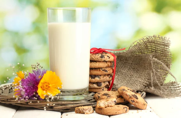 Glass of milk, chocolate chips cookies with red ribbon and wildflowers on wooden table on green background — Stock Photo, Image