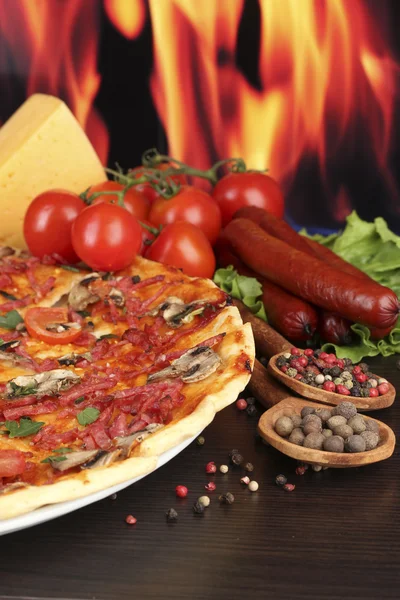 Pyszną pizzę, salami, pomidory i przyprawy na drewnianym stole na tle płomień — Zdjęcie stockowe