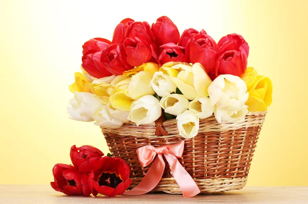 Beautiful tulips in basket on wooden table on yellow background — Stock Photo, Image