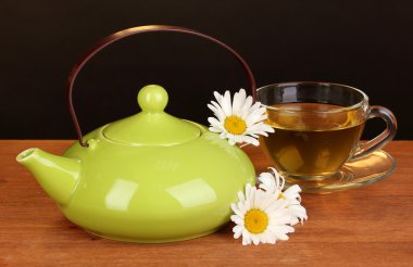 Teapot and cup with chamomile tea on wooden table on brown background clipart