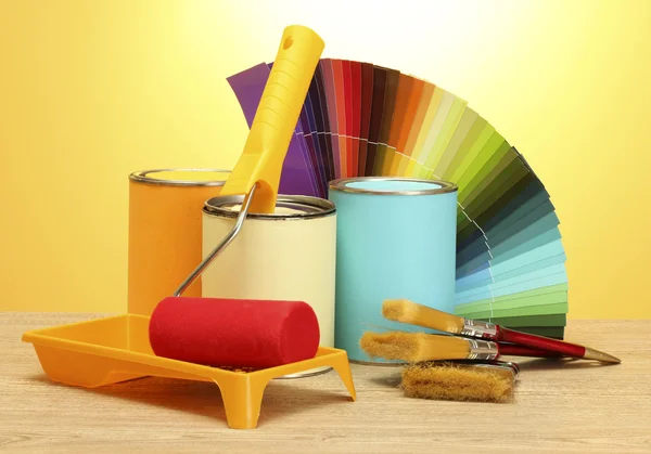 Жестяные банки с краской, ролики, кисти и яркая палитра цветов на деревянном столе на желтом фоне — стоковое фото