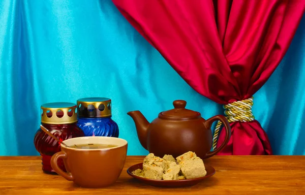 Чайник с чашкой и блюдцем со сладкой халвой на деревянном столе на фоне занавеса крупным планом — стоковое фото
