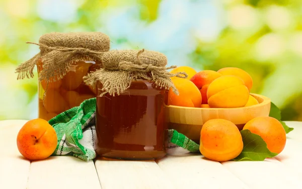 Canned apricots and jam in a jars and ripe apricots in bowl on wooden table on green background — Stock Photo, Image