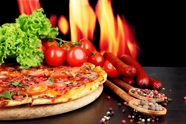 Delicious pizza, salami, tomatoes and spices on wooden table on flame background — Stock Photo, Image